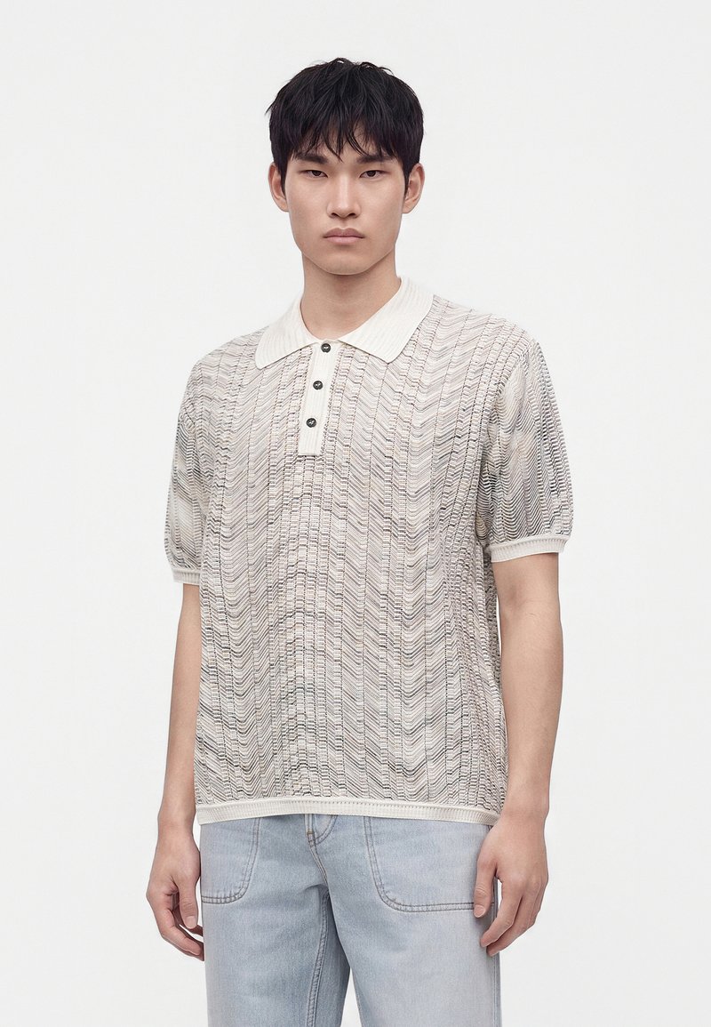 Jeune homme aux cheveux noirs courts portant un polo beige à manches courtes à motifs et un pantalon en jean bleu clair sur un fond uni.