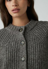Cárdigan gris de canalé hecho de un tejido de punto grueso, con un cuello redondeado y tres grandes botones redondos. La textura es cálida y acogedora.