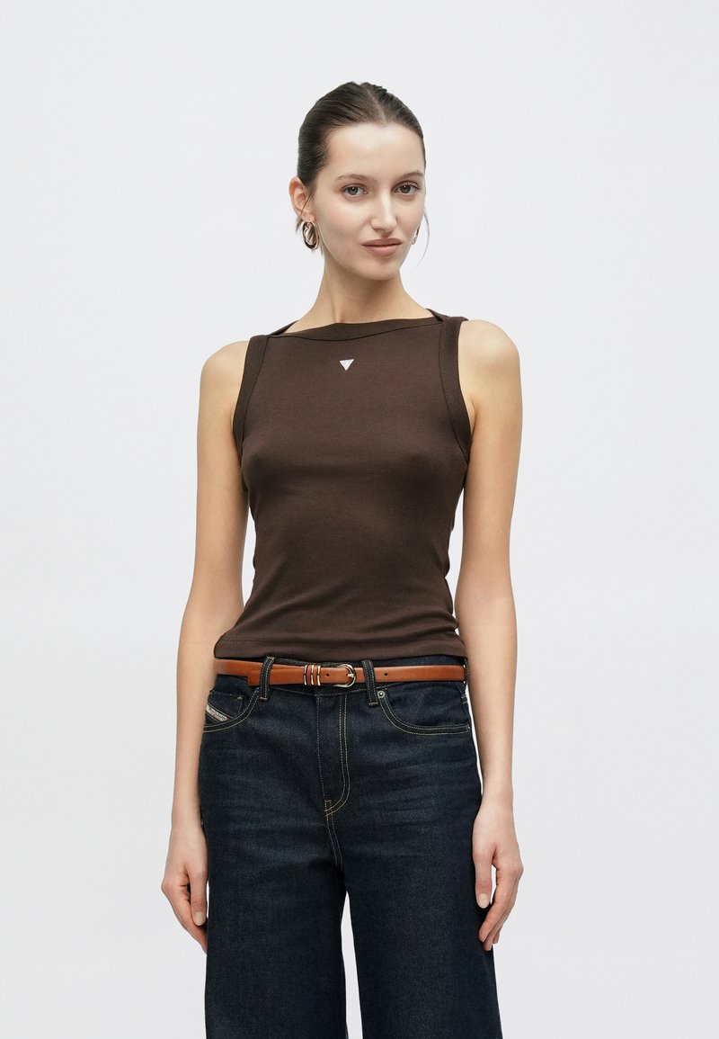 Rjava majica brez rokavov iz raztegljive tkanine, z diskretno trikotno nalepko na prsih, kombinirana s temno modrimi širokimi kavbojkami in rjavim pasom.