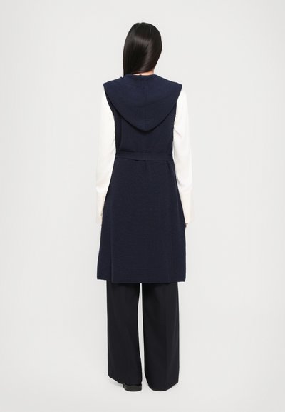 Mornarsko pleteno brezrokavnik s kapuco, z beljenim pasom, dolžino do kolen in teksturirano tkanino; v kombinaciji s širokimi črnimi hlačami.