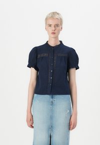 Marineblå kortermet bluse med blonder, rynket halsutskjæring og knappeåpning, kombinert med en lys blå denimskjørt med slitt.