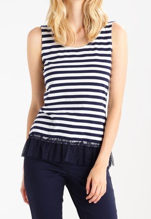 Vrouw draagt een mouwloze marineblauw-wit horizontaal gestreepte top met kanten rand bij de zoom en marineblauwe broek, staat met één hand ontspannen.