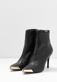 Czarne skórzane botki za kostkę z wysmukłymi noskami i złotymi metalowymi akcentami na czubku. Posiadają zamek błyskawiczny i wysoką szpilkę.