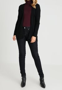 Černý kardigan přes vínově červený rolák, doplněný o tmavé skinny džíny a černé kotníkové boty. Hladká textura, otevřený přední design.