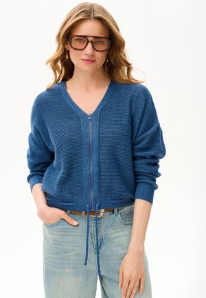 Kobieta nosząca duże okulary w oprawkach w kolorze muszli żółwia, niebieski sweter dziany z zamkiem z przodu oraz jasnoniebieskie dżinsy, stoi z jedną ręką w kieszeni.