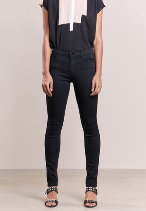 Osoba ubrana w obcisłe czarne dżinsy, czarno-beżową bluzkę oraz czarne sandały na otwartym palcu ze srebrnymi ćwiekami, stojąca na jednolitym tle.