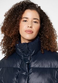 Navy puffer jacket featuring a high collar, glossy texture, and snap button closures. Curly hair frames the face. Simple background.