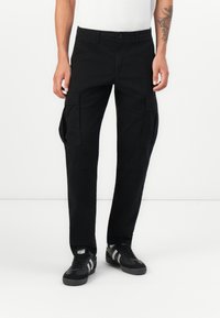 Sorte cargo bukser med flere sidelommer, et lige ben design og en robust tekstur, kombineret med sorte sneakers med hvide striber.