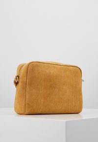 Pequeña bolsa cruzada rectangular de ante color beige con cierre de cremallera y asa metálica lateral, exhibida sobre una superficie blanca con fondo gris.