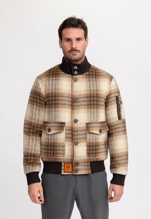 Mann trägt eine braun-beige karierte, geknöpfte Jacke mit gerippten Bündchen an Ärmeln, Kragen und Saum sowie graue Hose, steht vor weißem Hintergrund.