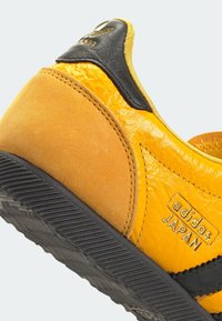 Žluté tenisky vyrobené z velurové a kožené materiály, s černými akcenty, texturovanými panely a detailem loga Adidas. Zakřivený design a gumová podrážka.