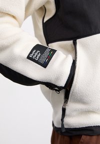Fleecejacke in Off-White mit schwarzen Akzenten, ausgestattet mit einem Reißverschluss und einem Label-Patch. Strukturierter Stoff und ein kontrastierendes Design.