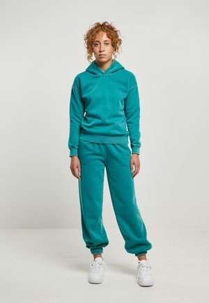 Felpa in cotone color teal con cappuccio regolabile e polsini a costine, abbinata a pantaloni della tuta tapered coordinati con elastico alle caviglie. Sneakers bianche.