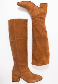 Un par de botas de ante marrón hasta la rodilla con tacones de bloque, una en posición vertical y la otra acostada en un fondo blanco.