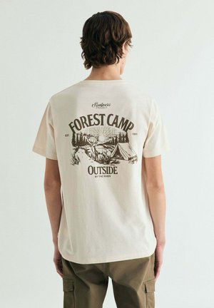 Persona con cabello corto y castaño que lleva una camiseta beige con la inscripción "Forest Camp" y una escena de campamento impresa en la espalda, de pie frente a una pared blanca.