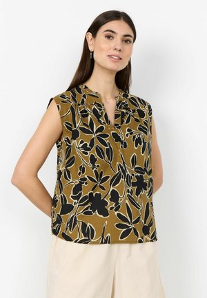 Femme aux longs cheveux noirs portant un haut marron sans manches avec un motif floral noir et un short crème, debout devant un fond blanc.