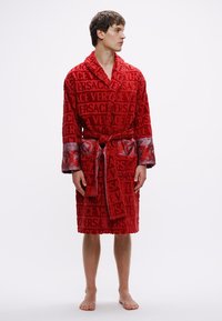 Červený župan z froté s výrazným vzorem loga „VERSACE“, kontrastními saténovými detaily na rukávech a zavazovacím páskem v pase.