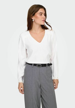 Femme portant une blouse blanche à manches longues avec des bords festonnés et un pantalon gris taille haute avec une ceinture noire, regardant de côté.