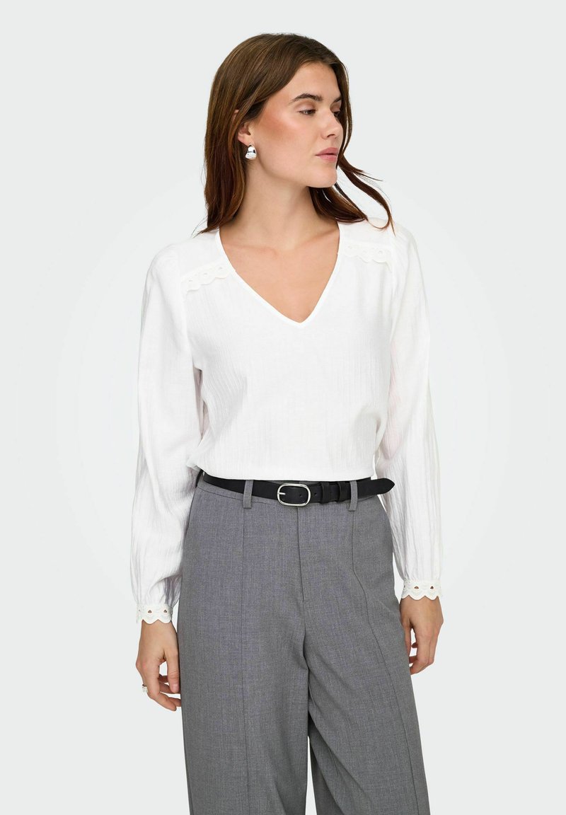 Femme portant une blouse blanche à manches longues avec des bords festonnés et un pantalon gris taille haute avec une ceinture noire, regardant de côté.