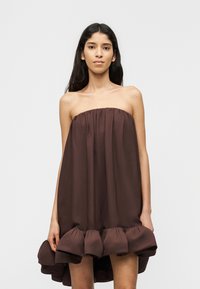 Γυναίκα που φοράει ιμάντα, φαρδύ καφέ φόρεμα με βολάν στο τελείωμα, στέκεται μπροστά σε ένα απλό ανοιχτό γκρι φόντο.