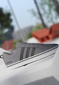 Zapatilla Adidas gris con rayas negras y suela blanca exhibida al aire libre sobre una superficie blanca, con árboles desenfocados y objetos naranjas en el fondo.