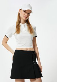Valkoinen cropped-polo paita mustalla reunuksella yhdistettynä mustaan A-linjaisiin hameeseen, jossa on kaksinkertaiset pyöreät metallirenkaat ja sivupleat.