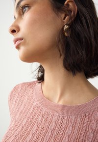 Lyserød strikket sweater med tekstureret mønster, ribstrikket halsudskæring og en tætsiddende pasform. Nærbillede, der fremhæver halsudskæring og stofdetaljer.