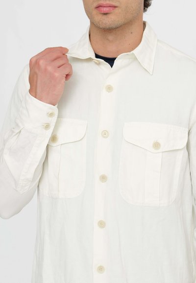 Camicia button-up di un colore crema chiaro, realizzata in un tessuto liscio. Presenta due tasche sul petto e un colletto classico, con polsini abbottonati sulle maniche.