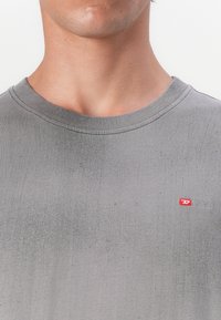 T-shirt cinza com decote redondo, apresentando uma textura subtil e um pequeno logotipo vermelho da Diesel no peito. Acabamento em tecido suave.