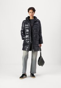 Glinsende mørkeblå pufferjakke med hette, frontglidelås og synlige sømmer. Modellen har på seg lyse denimjeans og svarte joggesko, og holder en veske.