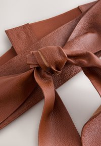 Brown leather belt featuring a textured finish, with a large tied knot detail and a smooth inner lining. Adjustable fabric strap included.