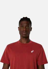 Červené sportovní tričko z lehkého materiálu. Má kulatý výstřih, krátké rukávy a bílé logo Asics na levém horním rohu.