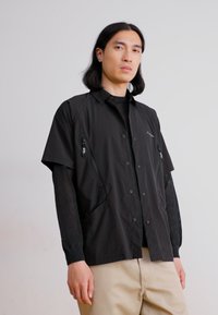 Black short-sleeve shirt with a button-up front, featuring zippered pockets and a lightweight fabric. Worn over a black long-sleeve shirt.