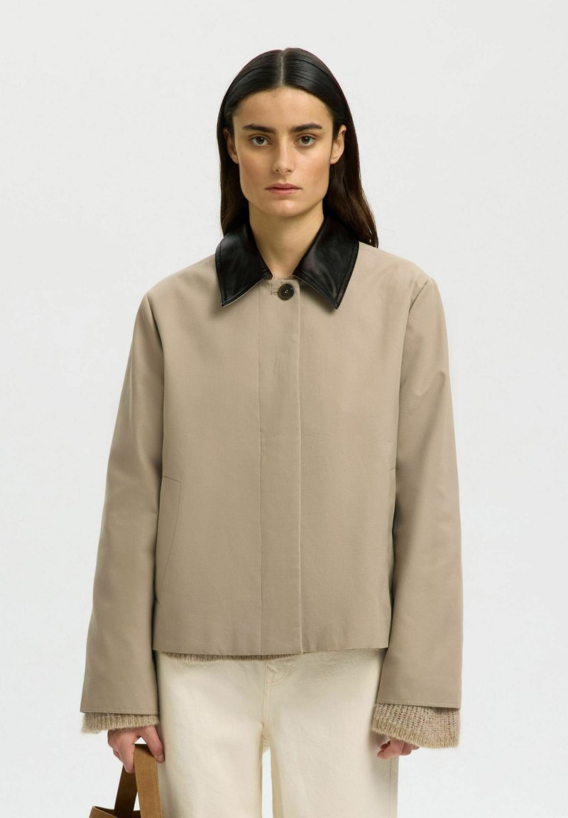 Femme aux cheveux noirs et raides, portant une veste beige avec un col noir, un pantalon crème, tenant un sac marron devant un fond clair uni.