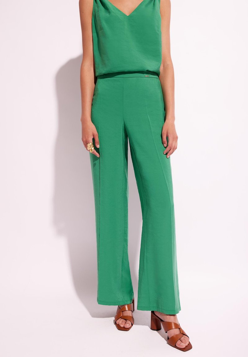 Zielona bezrękawnikowa bluzka w połączeniu z szerokimi zielonymi spodniami. Outfit charakteryzuje się gładką fakturą i luźnym krojem. Ubrane są brązowe sandały na obcasie.