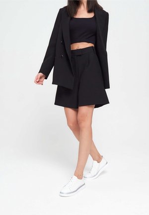 Mujer caminando con un blazer negro, un top corto negro, pantalones cortos negros y zapatillas blancas sobre un fondo blanco liso.