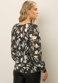 Svart blus med blommig tryck i kräm och beige, med långa ärmar, knäppning bak och en avslappnad passform. Slät, lättviktsmaterial.