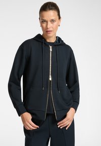 Donna con i capelli raccolti, che indossa una felpa con zip blu navy e pantaloni coordinati, in piedi con le mani vicino alle tasche su uno sfondo neutro.