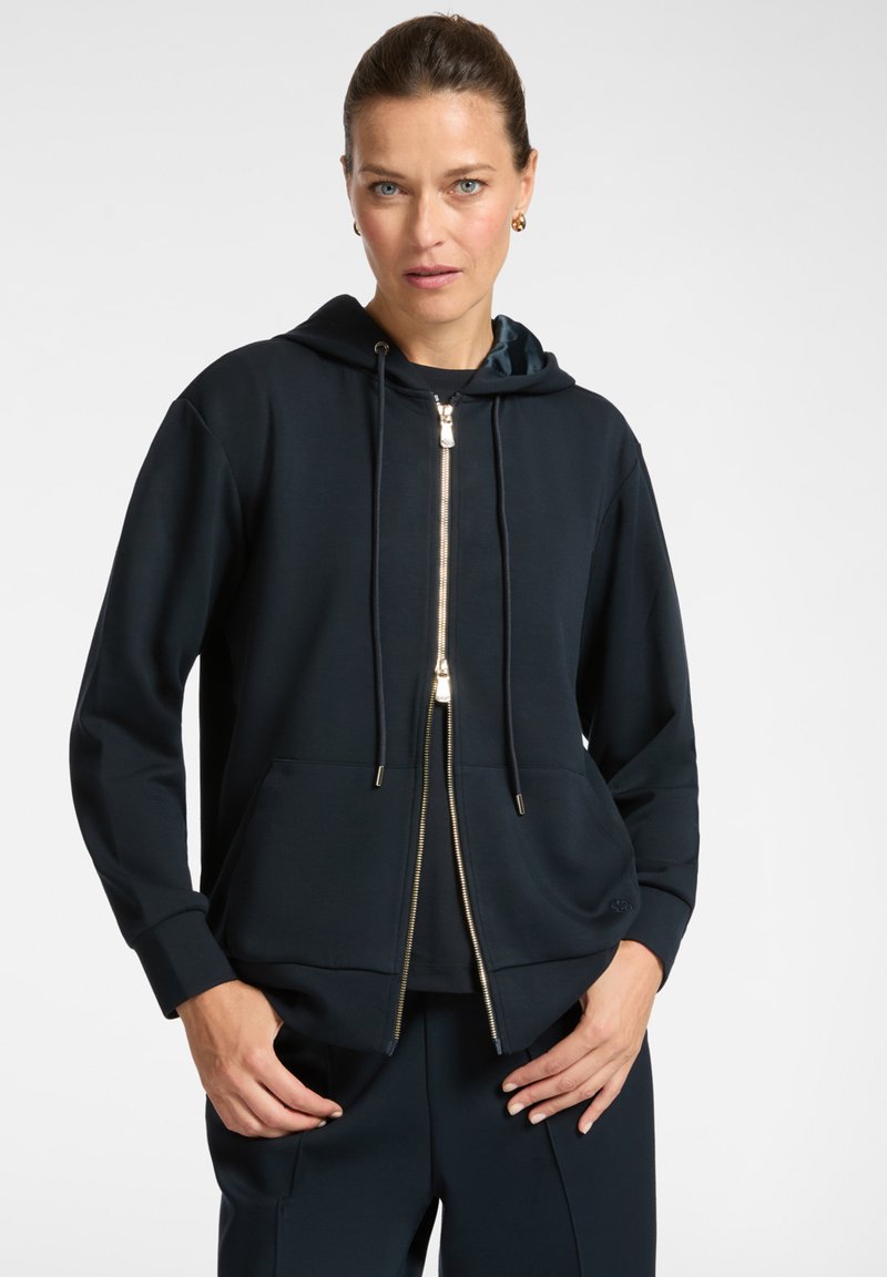 Donna con i capelli raccolti, che indossa una felpa con zip blu navy e pantaloni coordinati, in piedi con le mani vicino alle tasche su uno sfondo neutro.
