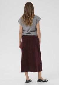 Donna con lunghi capelli castano chiaro, vestita con una maglietta grigia a maniche corte, una gonna midi in corduroy bordeaux e ballerine nere con borchie, in piedi con la schiena rivolta verso l'osservatore.