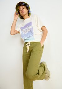 White graphic t-shirt with blue and green print. Olive green drawstring pants with pockets. Multicolored fuzzy hat. Light sneakers.