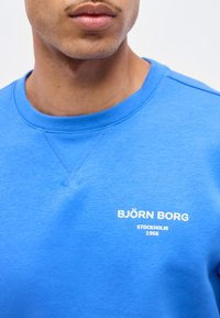 Άνδρας που φοράει μπλε φούτερ Björn Borg με το κείμενο "Stockholm 1956" στο στήθος, ορατός από τον λαιμό μέχρι το μέσο του κορμού.