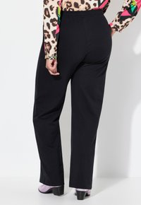 Zwarte, comfortabele broek met een elastische tailleband. Model draagt lavendelkleurige hakken. Achtergrond is eenvoudig wit.