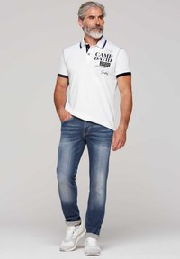 Polo bianco con bordi neri, con testo stampato; abbinato a jeans blu chiaro e sneakers grigie. Stile casual, vestibilità rilassata.
