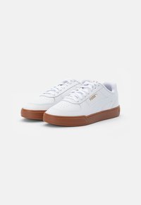 Zapatillas bajas blancas de Puma con suelas de goma marrón, área del dedo del pie perforada y logo dorado de Puma en el lateral y en la lengua.