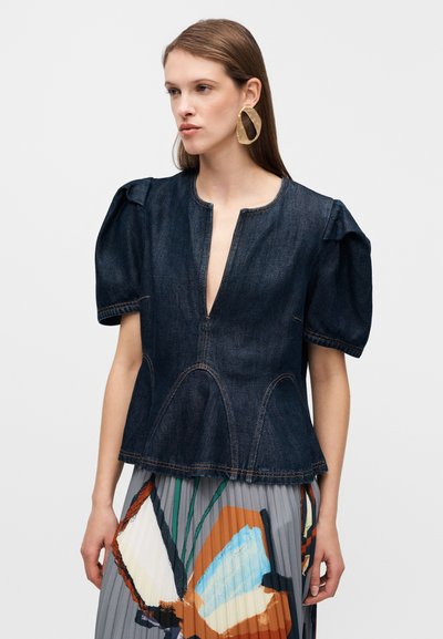 Haut en denim foncé avec manches bouffantes, décolleté profond, ourlet péplum et coutures contrastantes. Porté sur une jupe plissée à motifs.