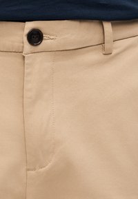 Μπεζ βαμβακερό παντελόνι με λεία υφή, ένα μαύρο κου μπί και ορατή ραφή. Διαθέτει μπροστινή τσέπη στην αριστερή πλευρά.