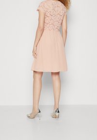 Robe rose pâle avec un corsage en dentelle, des manches courtes et une jupe évasée en chiffon. Comprend des chaussures à talons hauts argentées avec des accents décoratifs.