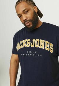 Námornícko modré tričko vyrobené z bavlny, s odvážnymi bielymi a zlatými nápismi. Dizajn obsahuje texty "ACK&JONES" a "EST. 90". Krátke rukávy.