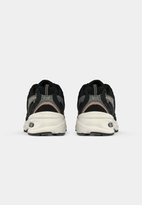 Um par de ténis atléticos pretos e bege mostrados pela parte de trás, apresentando solas brancas grossas e detalhes em tecido de malha.